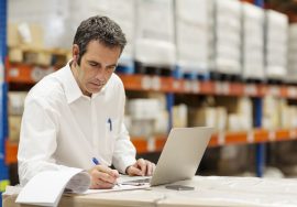 Despachante de aduana revisando documentación de importación junto a mercadería en depósito, con notebook abierta para gestión de despacho