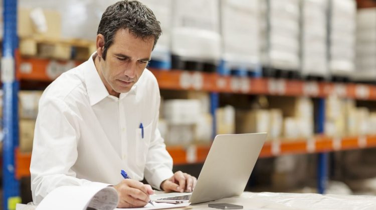 Despachante de aduana revisando documentación de importación junto a mercadería en depósito, con notebook abierta para gestión de despacho