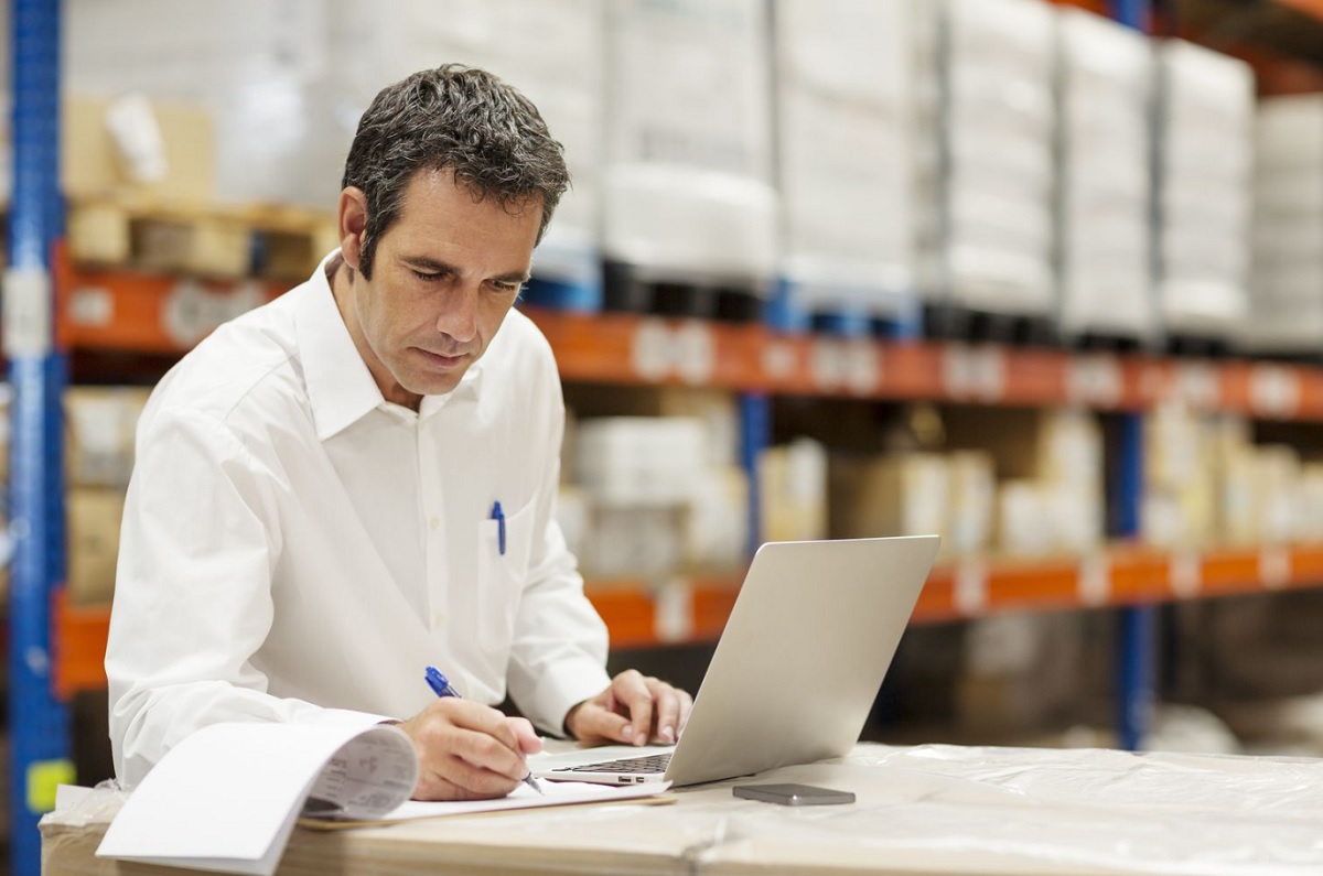 Despachante de aduana revisando documentación de importación junto a mercadería en depósito, con notebook abierta para gestión de despacho