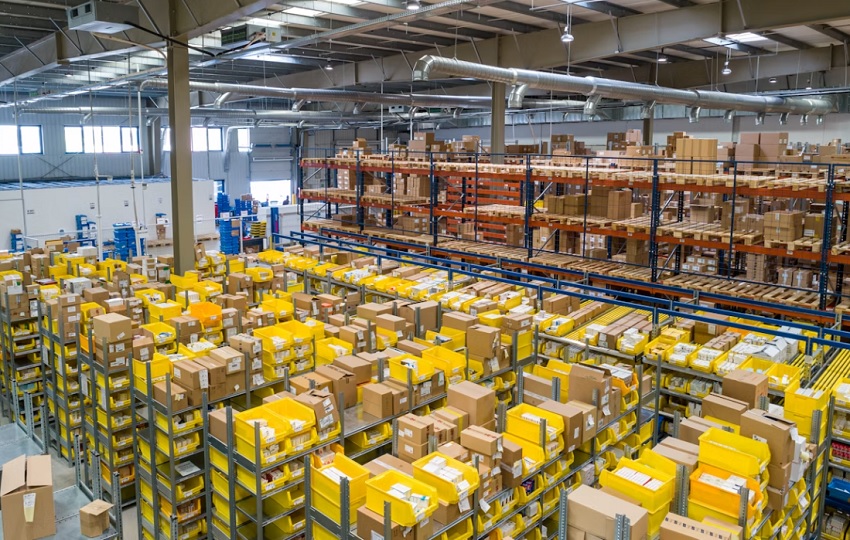 Warehouse interior with tall shelving, yellow bins and cardboard boxes organized on multiple racks under industrial ceiling ducts and lights.