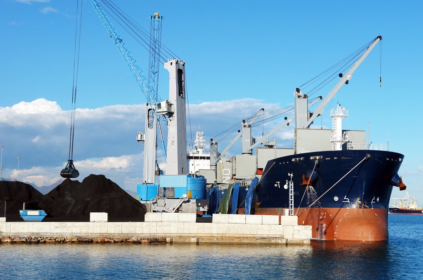 Cargo ship docked at a port with large cranes and a coal pile nearby, ready for loading.