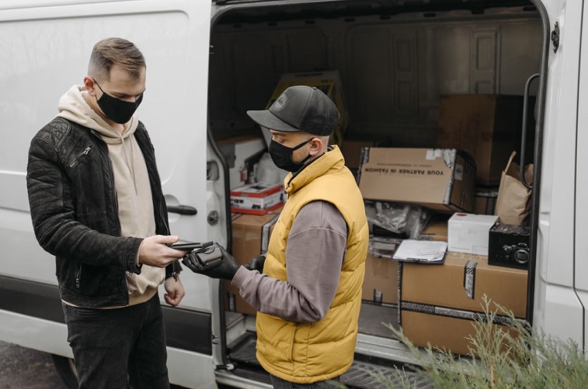 Delivery worker in a yellow vest and black mask hands a payment device to a customer by an open van filled with boxes, both wearing masks.